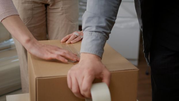 Couple packing up storage things in cardboard boxes, using adhesive sticky tape to wrap package for transportation. Preparing to move in household apartment bought on loan. Close up.