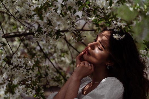 The sun shines on the girl's face near the flowering trees in the park. Portrait of attractive girl with curly hair in the garden