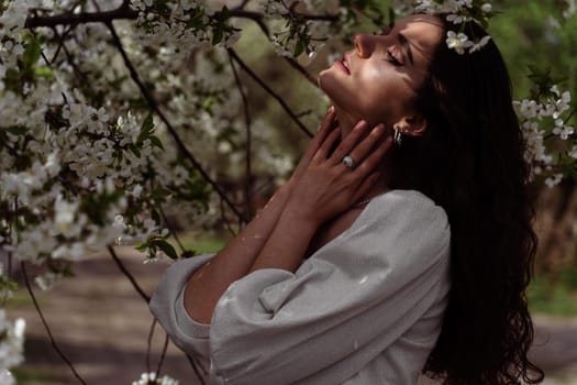 The sun shines on the girl's face near the flowering trees in the park. Portrait of attractive girl with curly hair in the garden