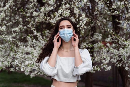 Girl in medical mask near white blooming trees in the park. Outdoor walking countryside at quarantine coronavirus covid-19 period. Spring lifestyle