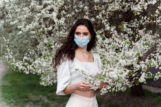 Girl in medical mask near white blooming trees in the park. Outdoor walking countryside at quarantine coronavirus covid-19 period. Spring lifestyle