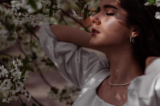 The sun shines on the girl's face near the flowering trees in the park. Portrait of attractive girl with curly hair in the garden