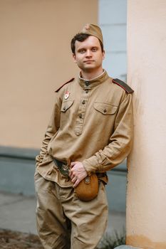 A man in a Soviet uniform from world war II. The soldier is standing against the yellow wall, holding his belt with his hands. Serious face