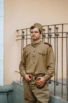 A man in a Soviet uniform from world war II. The soldier is standing against the yellow wall, holding his belt with his hands. Serious face