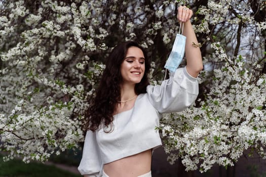 Girl holding medical mask after end of quarantine coronavirus covid-19 period. Model posing near white blooming trees in the garden