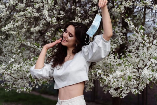 Girl holding medical mask after end of quarantine coronavirus covid-19 period. Model posing near white blooming trees in the garden