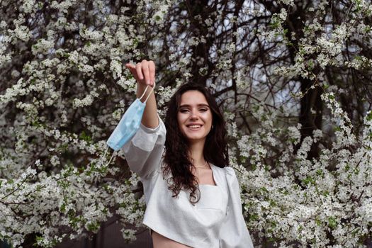 Girl holding medical mask after end of quarantine coronavirus covid-19 period. Model posing near white blooming trees in the garden