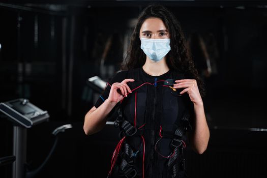 Girl in EMS suit and medical mask in gym. Protection from coronavirus covid-19. Sport training in electrical muscle stimulation suit at quarantine period