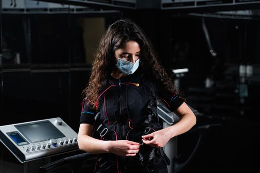 Girl in EMS suit and medical mask in gym. Protection from coronavirus covid-19. Sport training in electrical muscle stimulation suit at quarantine period