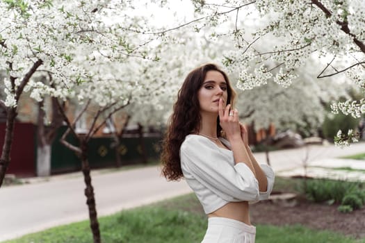 Spring lifestyle. Model posing near white blooming trees without mask outdoor countryside. Dreaming girl with curly hair