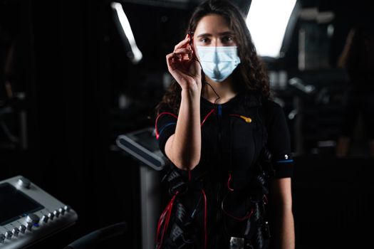 Girl in EMS suit and medical mask in gym. Protection from coronavirus covid-19. Sport training in electrical muscle stimulation suit at quarantine period