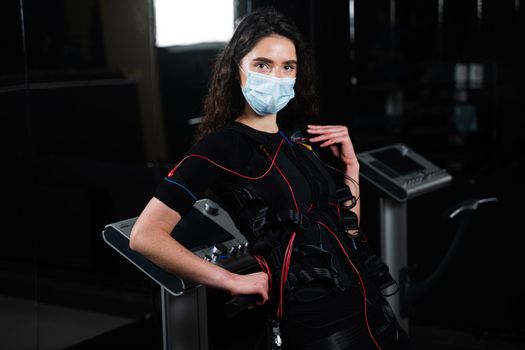 Girl in EMS suit and medical mask in gym. Protection from coronavirus covid-19. Sport training in electrical muscle stimulation suit at quarantine period