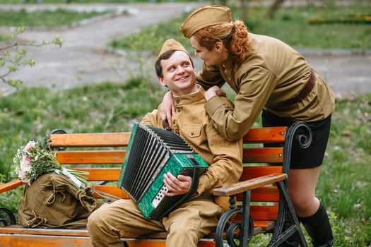 A soldier is sitting on a bench, playing an accordion. Behind is a girl military