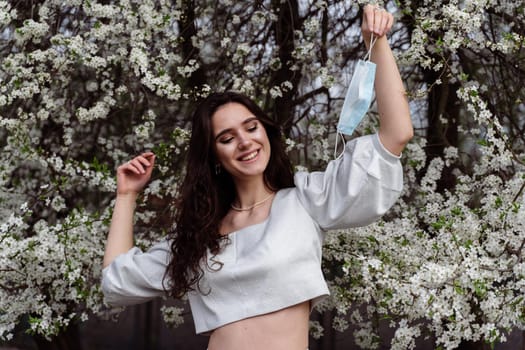 Girl holding medical mask after end of quarantine coronavirus covid-19 period. Model posing near white blooming trees in the garden