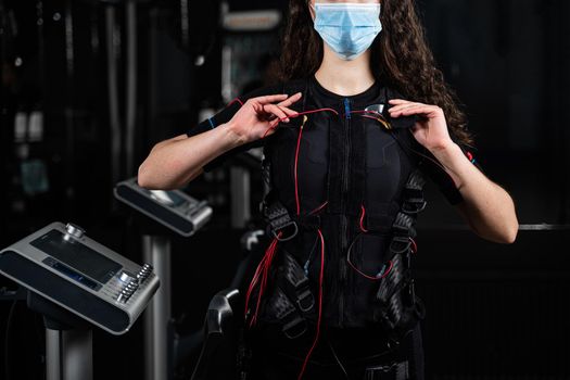 Girl in EMS suit and medical mask in gym. Protection from coronavirus covid-19. Sport training in electrical muscle stimulation suit at quarantine period