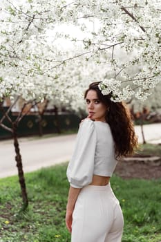 Spring lifestyle. Model posing near white blooming trees without mask outdoor countryside. Dreaming girl with curly hair