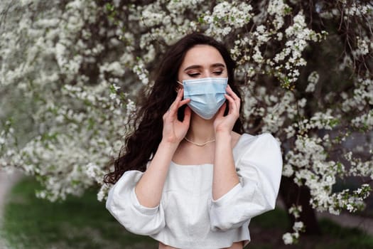 Girl in medical mask near white blooming trees in the park. Outdoor walking countryside at quarantine coronavirus covid-19 period. Spring lifestyle