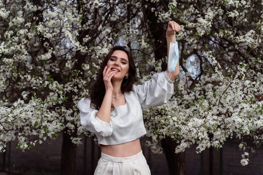 Girl holding medical mask after end of quarantine coronavirus covid-19 period. Model posing near white blooming trees in the garden