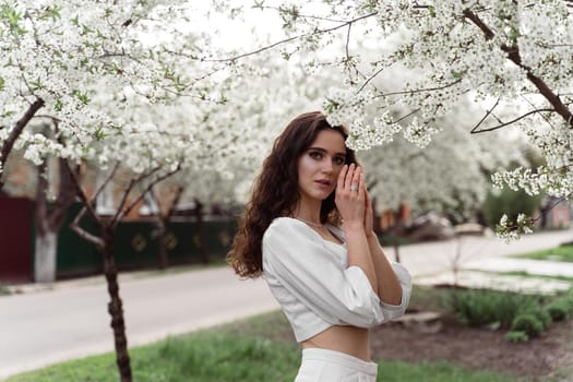 Spring lifestyle. Model posing near white blooming trees without mask outdoor countryside. Dreaming girl with curly hair