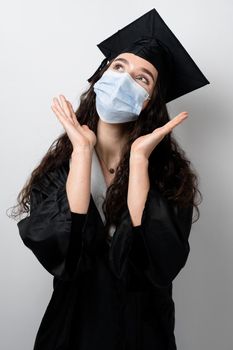 Surprised bachelor girl in medical mask at coronavirus covid-19 period. Master in graduation robe and cap on white background. Distance learning online. Study at home. Graduation from college.