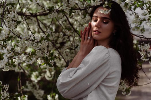 The sun shines on the girl's face near the flowering trees in the park. Portrait of attractive girl with curly hair in the garden