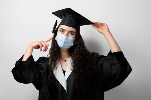 Graduate girl in medical mask at coronavirus covid-19 period. Woman with master degree in black graduation gown and cap. Distance learning online. Study at home. Graduation from college.