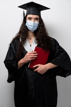 Student with book in graduation robe and cap in medical mask at coronavirus covid-19 period. Future leader of science. Academician young woman in black gown smiling.