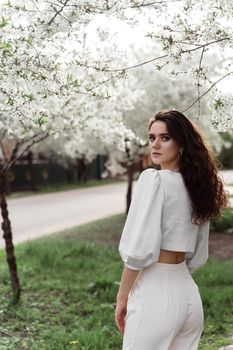 Spring lifestyle. Model posing near white blooming trees without mask outdoor countryside. Dreaming girl with curly hair