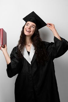 Student with book in graduation robe and cap ready to finish college. Future leader of science. Academician young woman in black gown smiling.