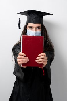 Student with book in graduation robe and cap in medical mask at coronavirus covid-19 period. Future leader of science. Academician young woman in black gown smiling.