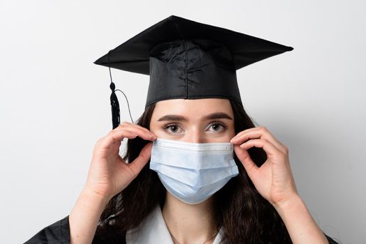 Graduate girl in medical mask at coronavirus covid-19 period. Woman with master degree in black graduation gown and cap. Distance learning online. Study at home. Graduation from college.