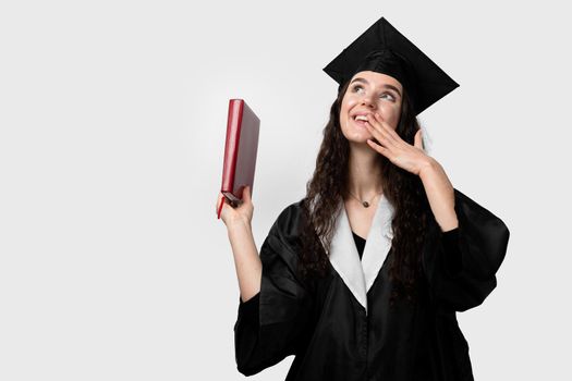 Student with book in graduation robe and cap ready to finish college. Future leader of science. Academician young woman in black gown smiling.