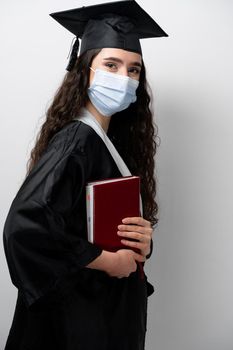 Student with book in graduation robe and cap in medical mask at coronavirus covid-19 period. Future leader of science. Academician young woman in black gown smiling.