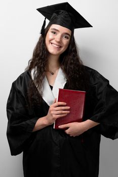 Student with book in graduation robe and cap ready to finish college. Future leader of science. Academician young woman in black gown smiling.