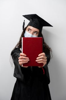 Student with book in graduation robe and cap in medical mask at coronavirus covid-19 period. Future leader of science. Academician young woman in black gown smiling.