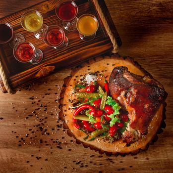 Pork knuckle with pickles, salad, vegetables, chili on a wooden board against the background of glasses with alcohol.