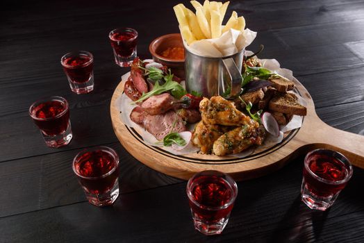Assortment of snacks from sausages, chicken wings, french fries, croutons, sauce on a wooden plate on a dark wooden table. Red alcohol in stacks around a plate of snacks.