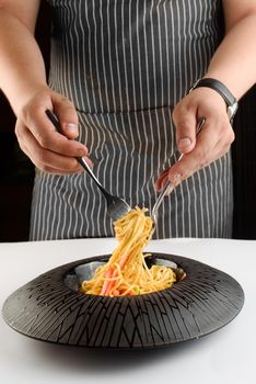 The chef stirs spaghetti with cheese, white sauce and cherry tomatoes with a fork and knife in a black plate on a white table.