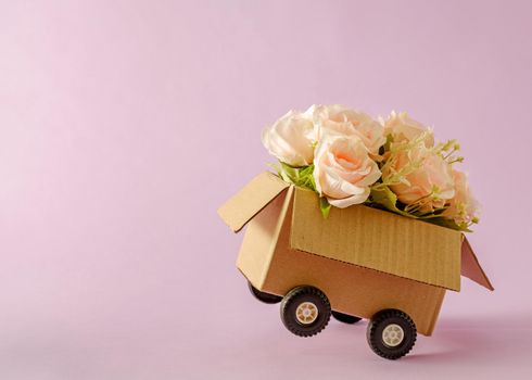 Cardboard box delivery container with truck wheels and bouquet of pink roses. Minimal concept of mail and congratulations with a holiday gift.