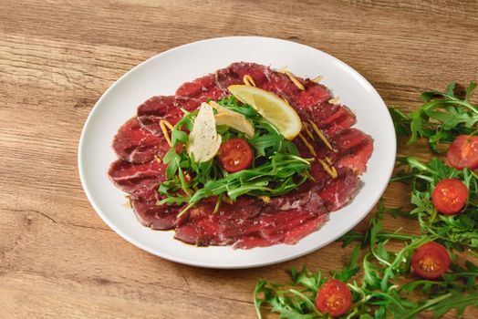 Carpaccio sliced meat with salad, tomatoes and lemon on a white plate on a wooden table.