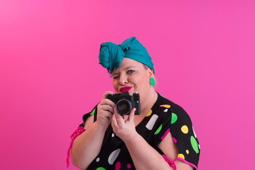 plus size smiling woman with funny emotional face expression with vintage camera in denim dress isolated on pink background, traveler on vacation, summer fashion style, excited tourist. High quality photo