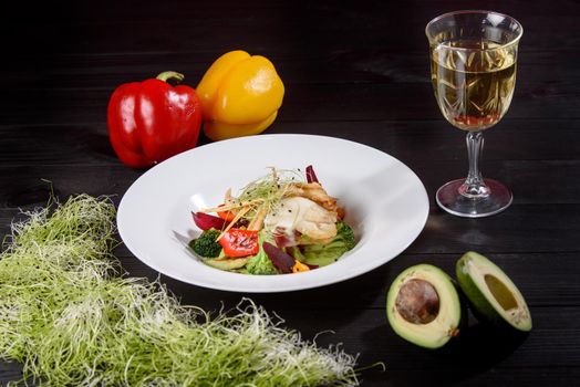 Chicken fillet with white sauce with grilled vegetables, garnished with microgreens on a white plate against the background of a glass of white wine, bell pepper and avocado on a wooden table.