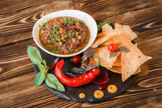 Mexican dish with meat, chili, tomato, corn with nachos in a white plate on a black slate platter, garnished with basil, blue onions and cherry tomatoes. Spicy Mexican dish on a wooden table.