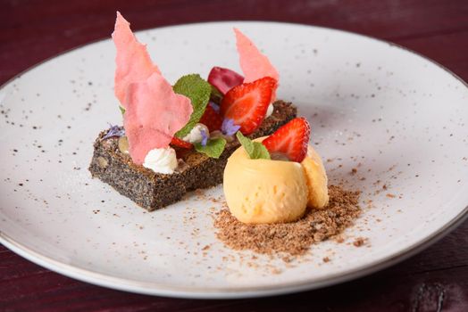 Poppyseed dessert with ice cream and biscuit crumbs, garnished with strawberry chocolate, strawberries, mint, cream and flowers on a white plate.