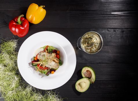 Chicken fillet with white sauce with grilled vegetables, garnished with microgreens on a white plate against the background of a glass of white wine, bell pepper and avocado on a wooden table.