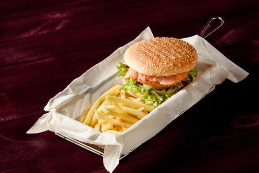 Burger with cutlet, bacon, salad, pickled cucumber and potato fries on parchment in a metal basket on a wooden background.