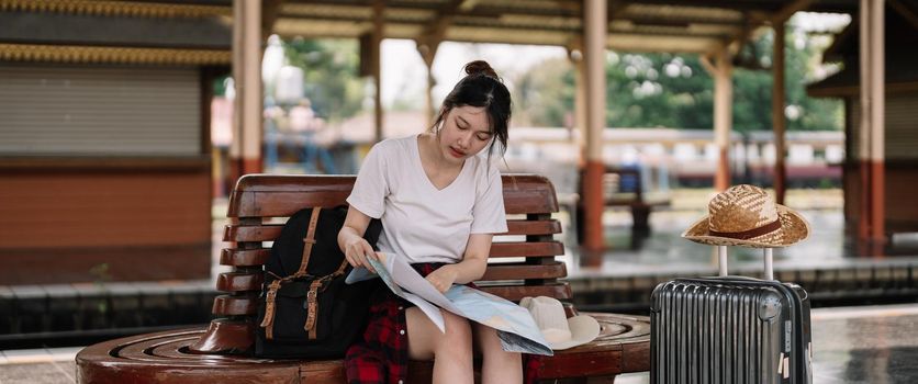 young asian woman traveler sitting with map choose where to travel and bag waiting for train at train station, summer vacation travel concept.