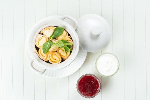 Beautiful serving of stuffed pancakes in a white ceramic pan with raspberry jam and sour cream garnished with mint on a white background. Nice breakfast and dessert. View from above