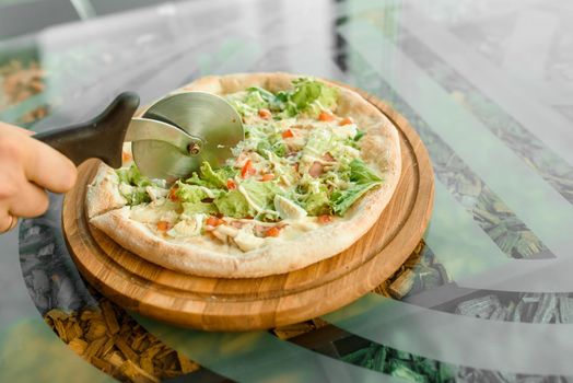 Pizza slicing using wheel knife pizza with ham, salami, tomatoes, salad and parmesan on a wooden board on a glass table