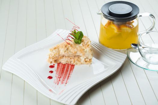 Napoleon cake decorated with a spiral of red chocolate, mint and berry jam on a white plate against a background of orange tea in a teapot and a transparent cup. Layered cake with pastry cream.